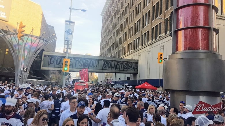 A crowd of people dressed in white shirts and other clothing and costumes, fill a downtown street. A sign in the background, across a skywalk, says Go Jets Go!!