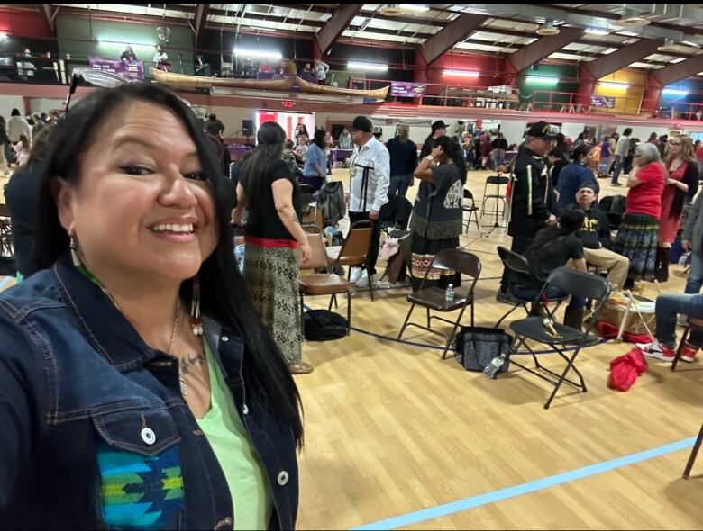 A woman with long dark hair and a jean jacket takes a selfie in a gym, many people standing behind her.