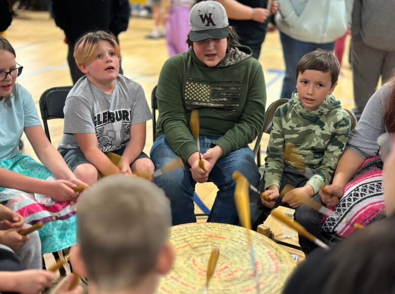 A group of children sit in a circle around a drum, each one holding a drumstick. 