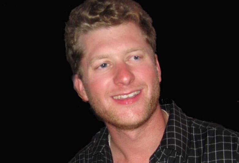 A man with wavy blonde hair wears a dark checkered shirt and smiles for a photo.