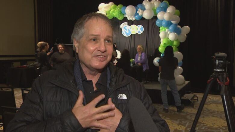 A man in black jacket smiles as he stands in a hotel conference room, with his hands over his heart.