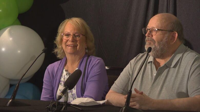 A man in a grey collared T-shirt and a smiling woman in a purple sweater and white floral shirt sit at a table in front of microphone.