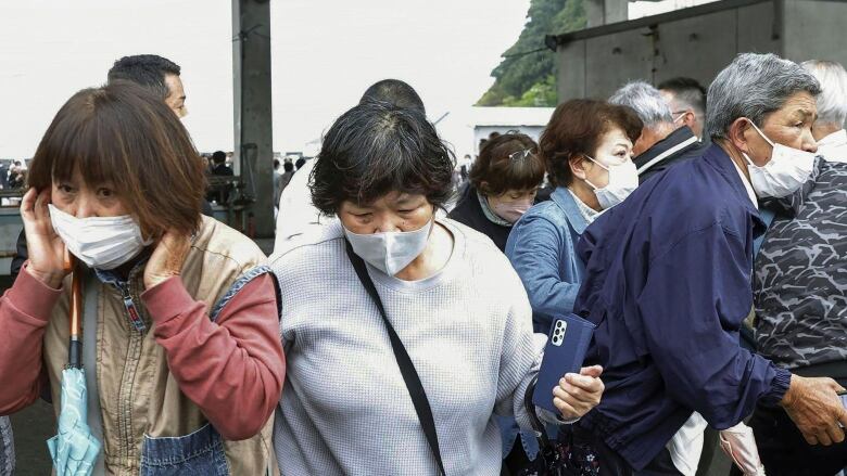 A group of elderly people wearing surgical masks is seen outside. One woman holds her hands to her ears.