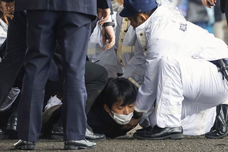 A man wearing a surgical mask is prone on the ground with several people standing or kneeling over him.