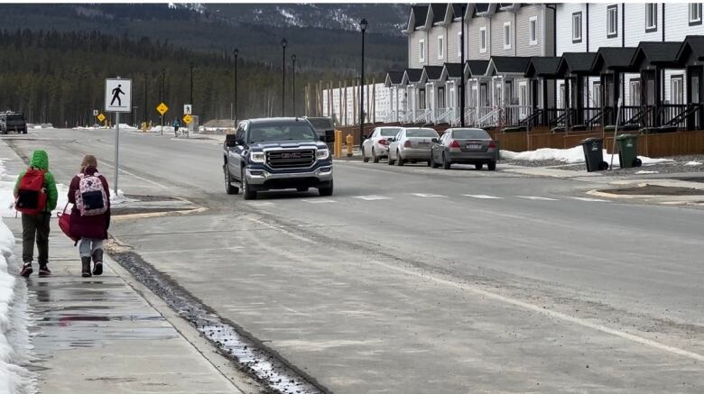 Photo shows two kids walking on the sidewalk, with a car driving by houses. 