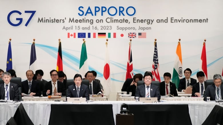 A group of men in suits sit at a long table with microphones in front of a sign.