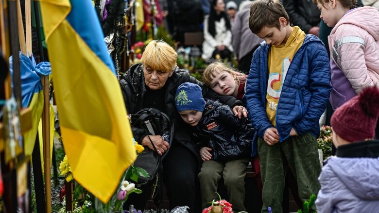 A woman and children gather solemnly by a grave with a Ukrainian flag and flowers.