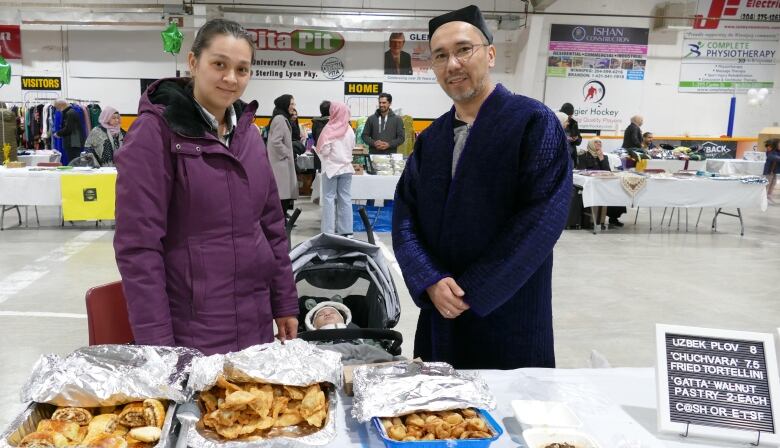 Two people are pictured standing in front of a table of food.