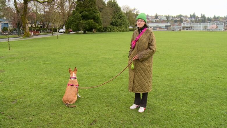 A woman with a toque on and wearing a long winter coat, looks one way, while a ginger-coloured dog on a lease looks the other at a park in Vancouver.