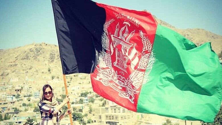 A woman in sunglasses and a head covering stands next to the Afghanistan flag, which is black and red and green, with mountains in the background.