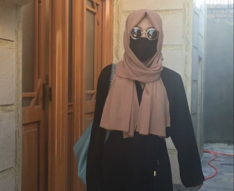 A woman stands in an alley covered completely in black robes, a head covering and sunglasses.