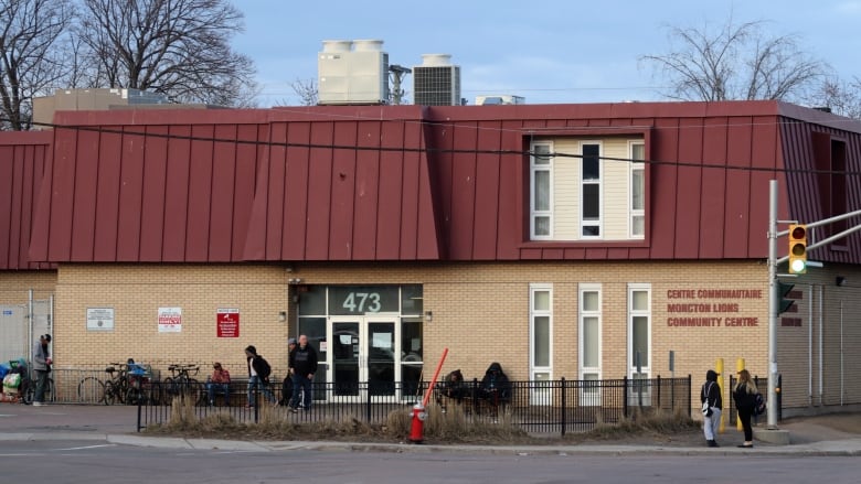 Several people standing outside a two-storey building made of yellow bricks a red metal roof.