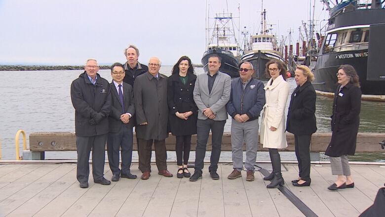 Ten people stand in front of fishing vessels on a dock on a blustery day.