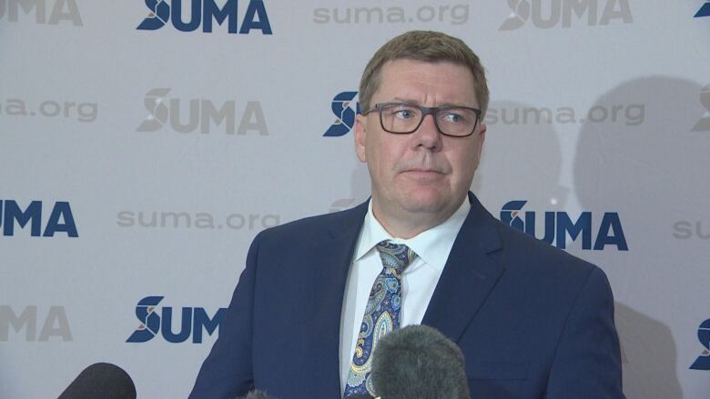 A white man with brown hair and glasses is wearing a three piece suit. The jacket is navy blue, his collared shirt is white and he has a tie with a multi-colour design on it.