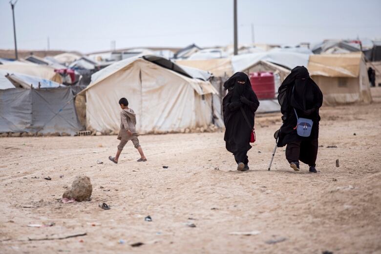 2 people in black burquas walk in the foreground of a tent encampment. A barefoot child is in the background.