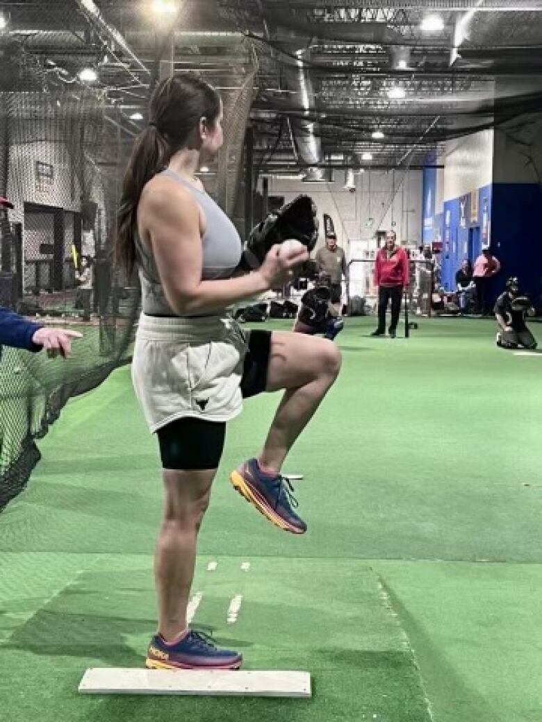 A woman holds a baseball and prepares to pitch inside a training facility.