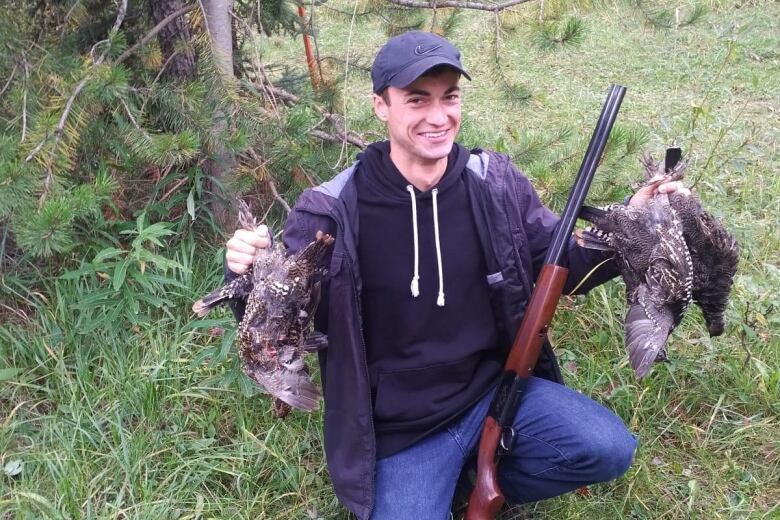 A man holds two dead birds with a riffle leaning against his knee.