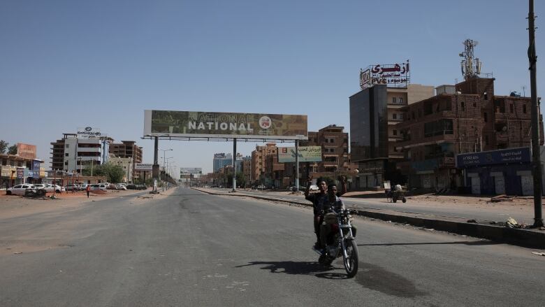 Two people on a motorbike drive down a deserted street with a billboard in the distance behind them.