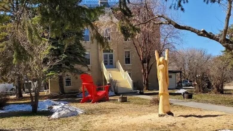 A wooden sculpture of an angel stands in front of a building and a large red muskoka chair. 