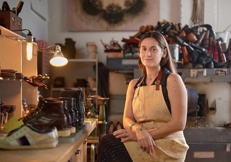 A woman sits cross-legged on a stool. She's surrounded my leather shoes in a studio workspace.