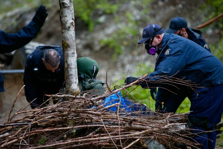 Police officers attend to a person surrounded by branches in a forest.