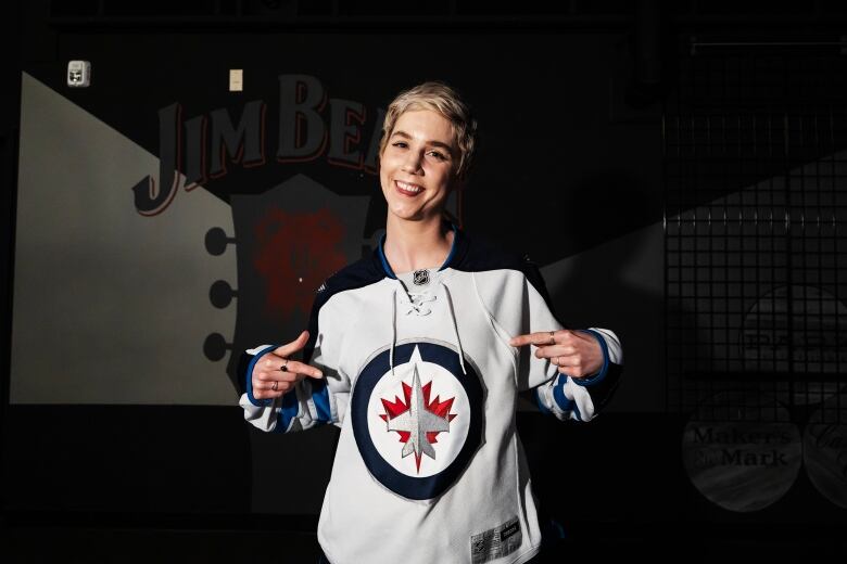 Smiling woman in Winnipeg Jets jersey points to the logo on her chest with both hands.