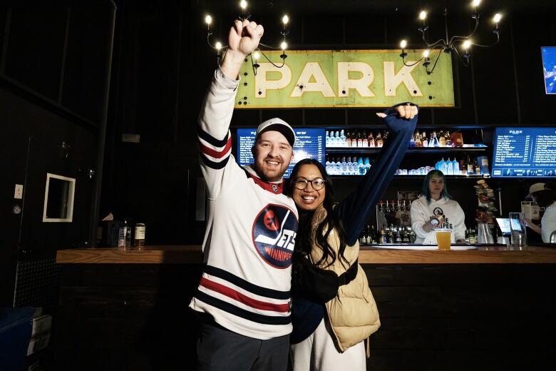 Man in Winnipeg Jets jersey and woman in tan jacket hug side-by-side with their outside arms raised in celebration.