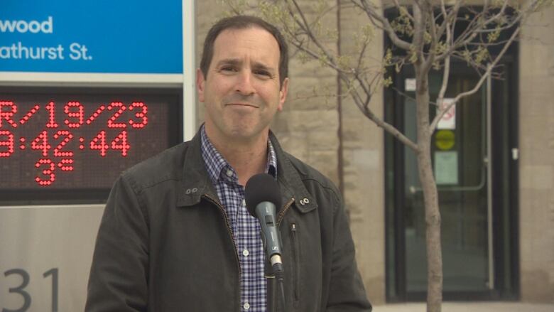 A man in a jacket and checked shirt smiles behind a microphone.