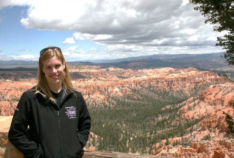 A woman in a black jacket stands above a mountainous landscape with white and grey clouds moving across a blue sky. 