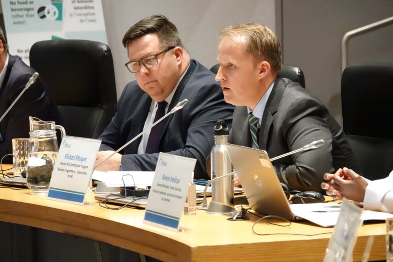 A man in a suit sits at a table and speaks into a microphone while another man in a suit beside him listens.