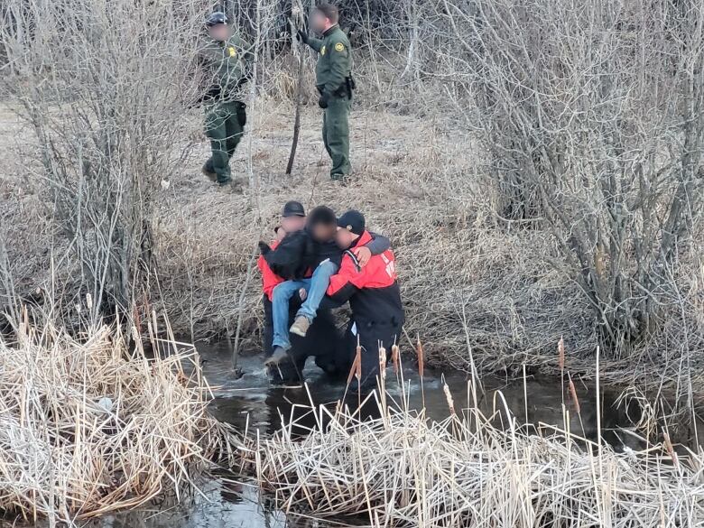 Men in drysuits carry a man in jeand and a hoodie across a stream.