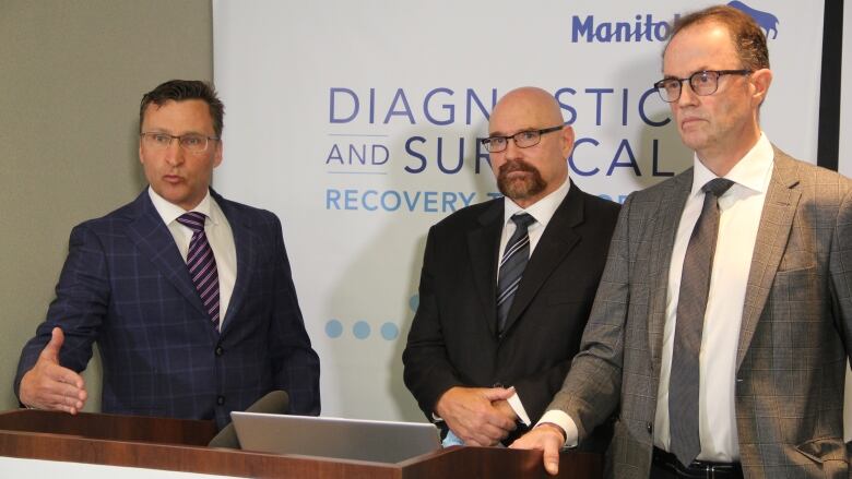 Three men stand and talk to the media behind a podium.