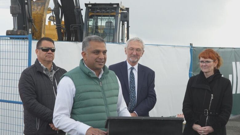 A South Asian man speaks at a podium with two men and a woman behind him.