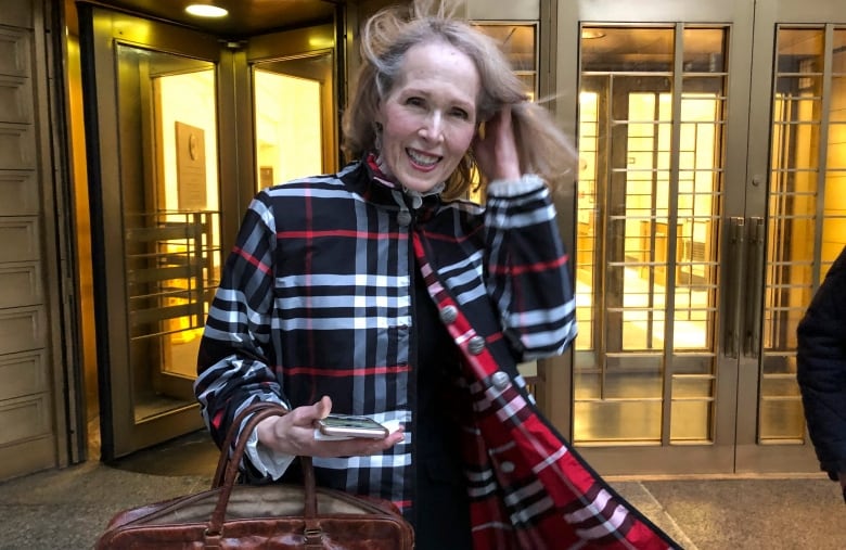 A woman in a coat brushes her hair back while walking outside a building entrance.