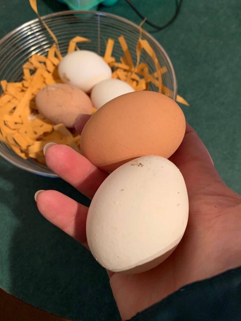 A hand is holding tow eggs - one brown, one white. The background shows a bowl with three more eggs.