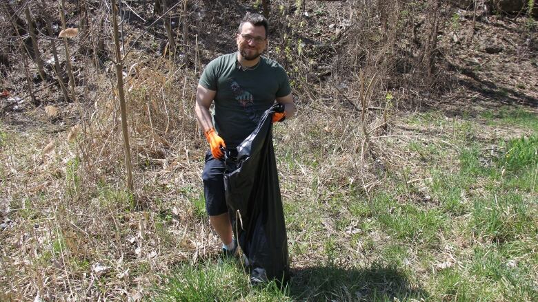 A man in a green shirt stands with gloves on holding a black trash bag 