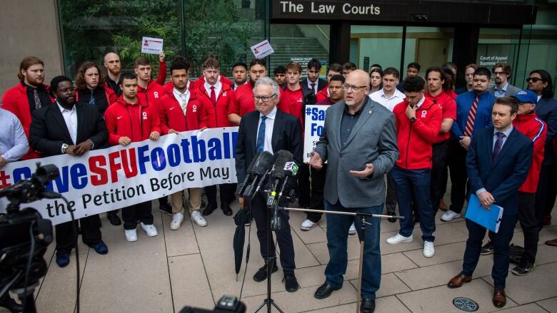 Alumni and supporters of the Simon Fraser Football program are pictured outside of the courthouse in Vancouver, on April 13, 2023. Many wear red jackets, and some hold a large sign that says Save SFU Football.