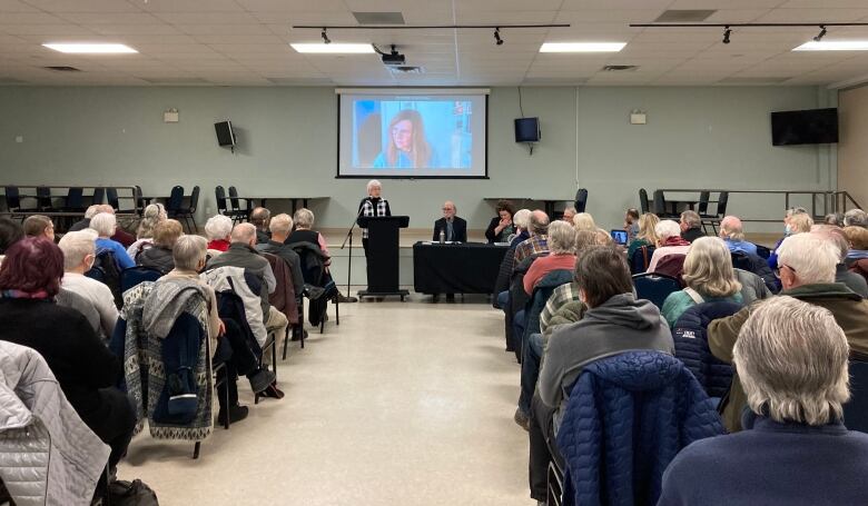 A large crowd is seen seated while a keynote speaker talks in front of a podium in a community centre room.