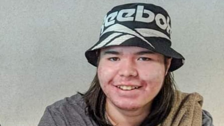 A young man with long hair and hands clasped in front of him smiles at the camera.