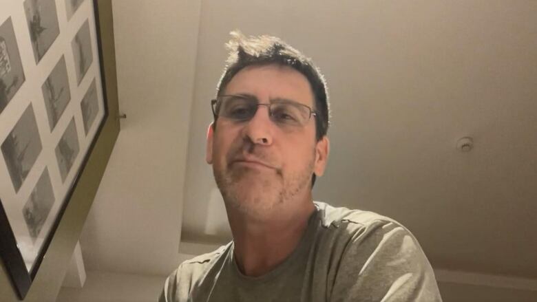 A man with glasses is seen looking down at a camera in a hotel room with a framed picture on the wall. 