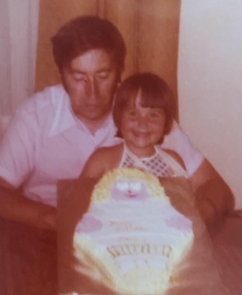 A young child smiles. She sits on a middle-aged man's lap. In front of them, on a table, is a cake.