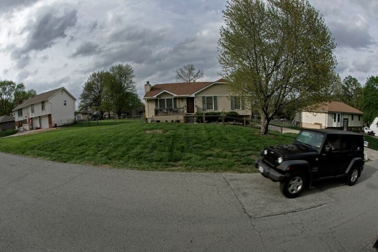 A jeep drives past a house, lawn and tree on a residential street.