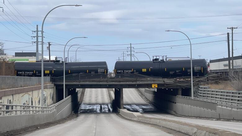 A black tanker from a train leans out from a concrete bridge above a street.