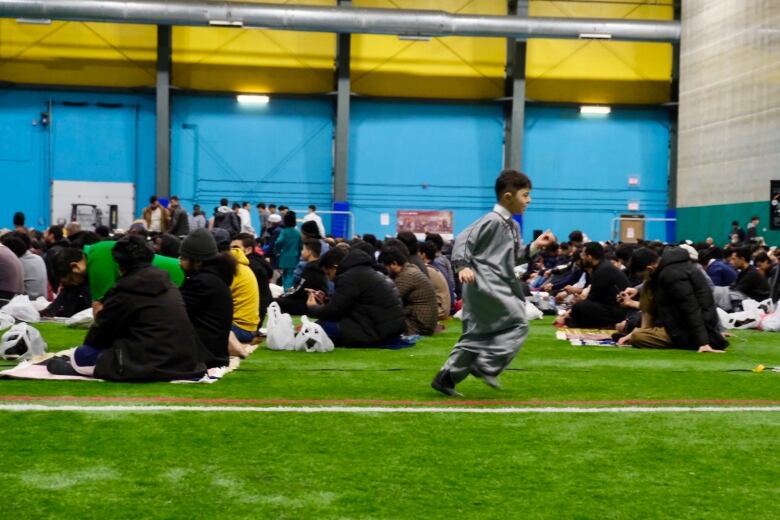 A boy runs past a group of people sitting on carpets on the ground at an indoor facility for Eid celebrations in St. John's 