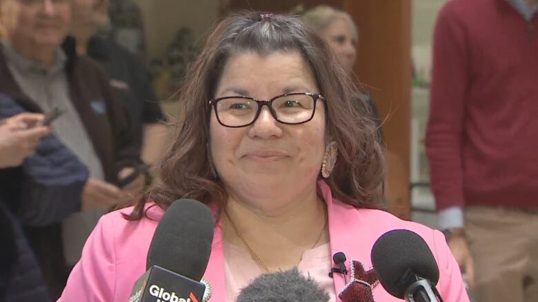 A woman with long brown hair and glasses wears a pink blazer with a pink shirt. She is standing at a podium with microphones.