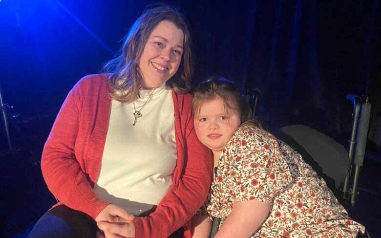 A mother and her daughter sit in front of a black curtain. There's a shining blue light in the top left corner, and the daughter is leaning on her mother's arm.