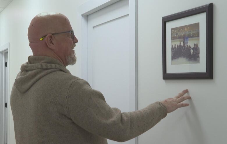 Para hockey player Eric Payne smiles looking at an old team photo on the wall. He is wearing a beige sweater. 