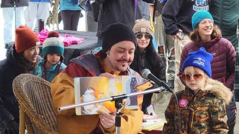 A man in yellow jacket sits behind a microphone with children and women around him.