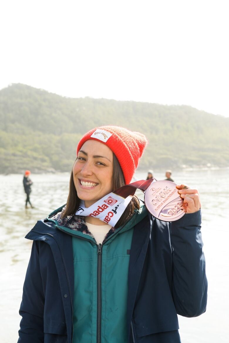 A woman in a bright red toque smiles and holds up a large bronze medal.