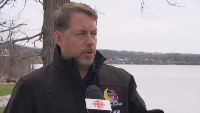 A man in a black jacket speaks to a reporter outdoors on a cloudy day.